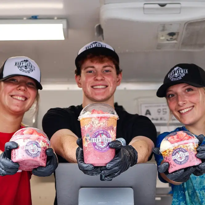 Three Buoy Bowls employees smiling and handing açaí bowls in the window of the purple truck